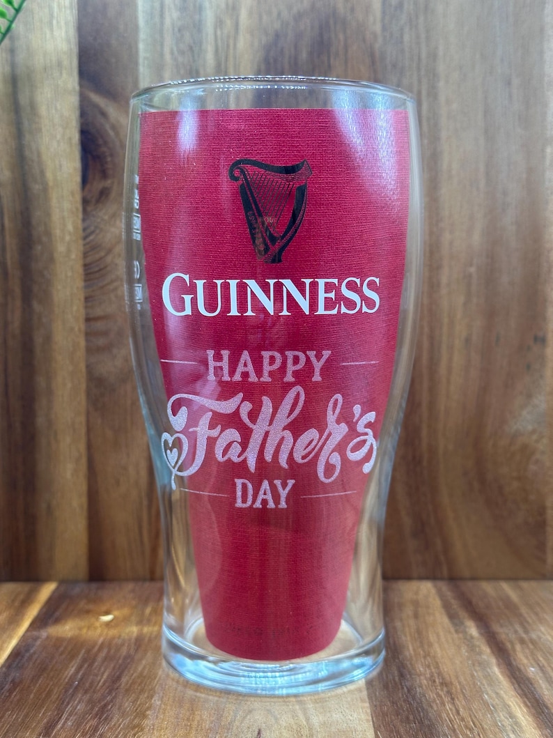 May include: A clear pint glass with a red insert featuring the Guinness logo and the words "Happy Father's Day" in white script. The glass has a curved shape and is designed for beer.
