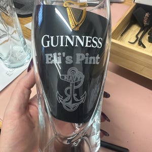 May include: A clear glass pint glass with a black background and white text that reads "GUINNESS Eli's Pint". A gold harp logo with the text "ESTD 1759" is at the top of the glass. A silver anchor with the word "NAVY" is at the bottom of the glass.