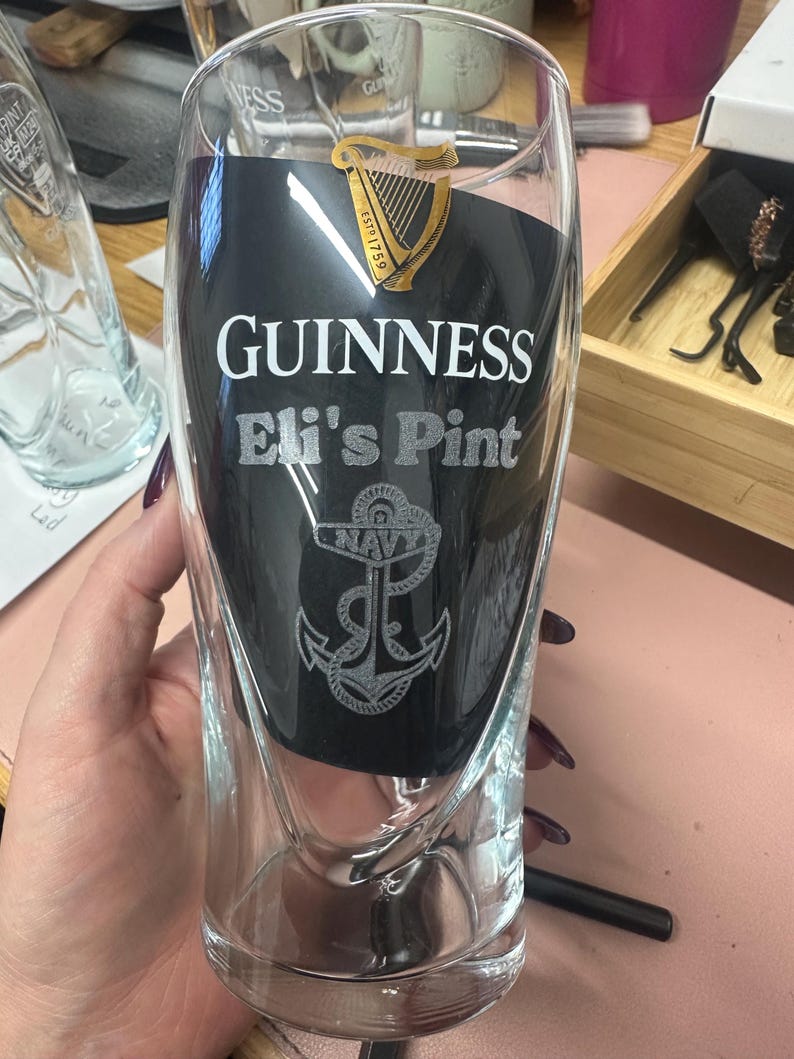 May include: A clear glass pint glass with a black background and white text that reads "GUINNESS Eli's Pint". A gold harp logo with the text "ESTD 1759" is at the top of the glass. A silver anchor with the word "NAVY" is at the bottom of the glass.