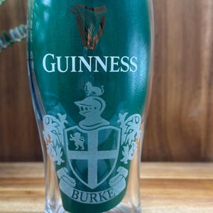 May include: A clear pint glass with the Guinness logo and the name "Burke" etched on it. The glass has a green background and a classic design, perfect for enjoying a pint of stout.