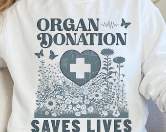 Puede incluir: Sudadera blanca con un estampado gráfico de un corazón con una cruz en su interior, rodeado de flores. El texto "ORGAN DONATION" está por encima del corazón y "SAVES LIVES" está por debajo del corazón.