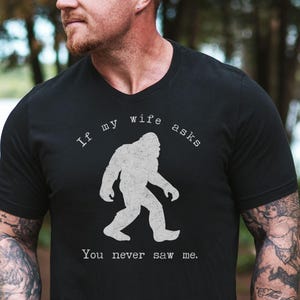 A black Bigfoot tee with a distressed Sasquatch silhouette in the center. The curved text reads If my wife asks and the lower text reads You never saw me. Funny camping T shirt theme with outdoorsy hiking humor and a simple centered design.
