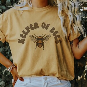 May include: A mustard yellow t-shirt with a brown bee graphic and the text "Keeper of Bees" in a retro font.