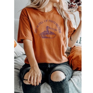 Optimistic Pasta Shirt in the color yam. A picture of a fork in a bowl of pasta with the text above that reads life is full of pasta-bilities.