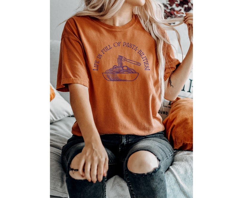 Optimistic Pasta Shirt in the color yam. A picture of a fork in a bowl of pasta with the text above that reads life is full of pasta-bilities.