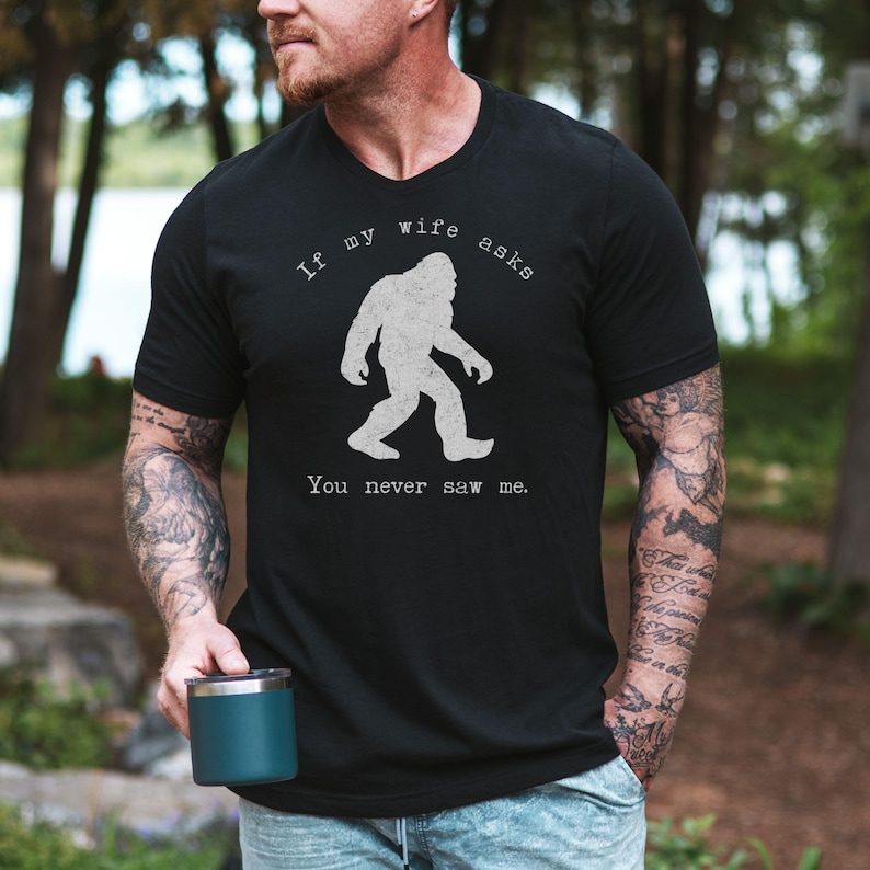A black Bigfoot tee with a distressed Sasquatch silhouette in the center. The curved text reads If my wife asks and the lower text reads You never saw me. Funny camping T shirt theme with outdoorsy hiking humor and a simple centered design.