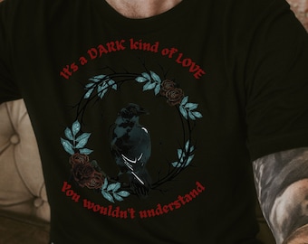 Op de afbeelding: Een zwart T-shirt met korte mouwen en een afbeelding van een kraai in een cirkelvormige krans van donkere rozen en blauwgroene bladeren. De rode tekst boven en onder de krans luidt, It's a DARK kind of LOVE You wouldn't understand.