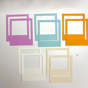 Op de afbeelding: Een verzameling kleurrijke papieren fotolijsten in tinten paars, blauw, oranje, wit en crème. De lijsten zijn gerangschikt op een wit oppervlak, met een houten kist op de achtergrond.