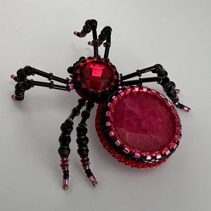 Könnte beinhalten: Eine schwarze Perlenspinne mit einem roten Strassstein-Körper und einem rosafarbenen und roten Perlenkreis.
