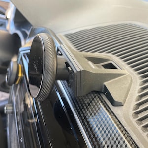 May include: Black car phone mount with a carbon fiber textured magnetic phone holder. The mount is attached to the dashboard. The phone holder is round and has a textured surface. The dashboard has a geometric pattern.