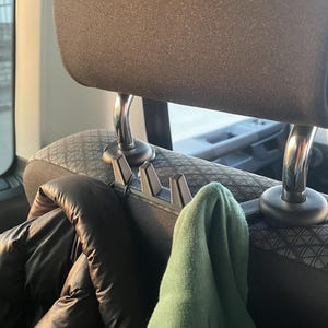 Könnte beinhalten: Eine Auto-Kopfstütze mit einem schwarzen Hakenaufsatz, der eine schwarze Jacke und ein grünes Kleidungsstück hält. Die Kopfstütze ist dunkelgrau und der Sitz hat ein Muster. Die Haken sind zum Aufhängen von Gegenständen konzipiert.