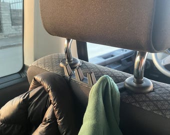 May include: A black car headrest with a hook attachment holding a black jacket and a green garment. The headrest is gray and black, with silver metal supports. The hooks are designed to hang items.