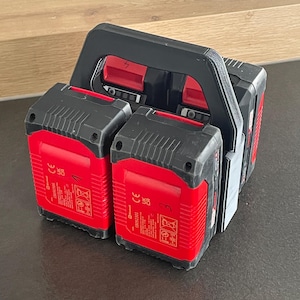 Könnte beinhalten: Ein schwarzes und rotes Ladegerät für Elektrowerkzeugbatterien mit einem schwarzen Griff. Zwei rote Batterien sind am Ladegerät befestigt. Die Batterien haben schwarze Oberteile und rote Etiketten mit weißem Text und Symbolen. Das Ladegerät steht auf einer dunklen Oberfläche.