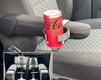 May include: A red Coca-Cola Zero Sugar can sits in a gray car cup holder attached to a car armrest. The armrest is black and gray. The image also shows a car console with two wine glass holders.