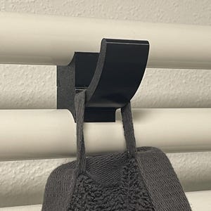 Puede incluir: Un gancho de radiador de metal negro está montado en un radiador blanco. Una bolsa de punto gris cuelga del gancho. El gancho tiene un diseño curvo y está fijado al radiador con una base plana.