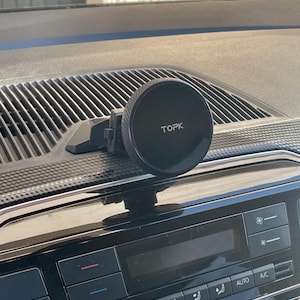 Może przedstawiać: Czarny uchwyt samochodowy na telefon z napisem "TOPK" na okrągłym uchwycie magnetycznym. Uchwyt jest przymocowany do deski rozdzielczej samochodu z widocznymi otworami wentylacyjnymi i przyciskami sterowania.