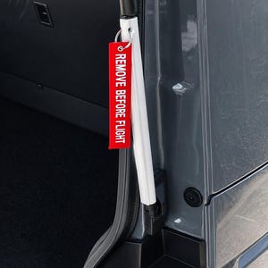 Può includere: Un'etichetta rossa "REMOVE BEFORE FLIGHT" è appesa al telaio della porta di un veicolo. L'etichetta ha testo bianco. La porta del veicolo è grigia e l'interno è nero.