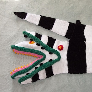 Pode incluir: Um cobertor de crochê listrado em preto e branco com uma cabeça de crocodilo verde e rosa em uma extremidade. A cabeça de crocodilo tem pontos pretos e um olho amarelo.