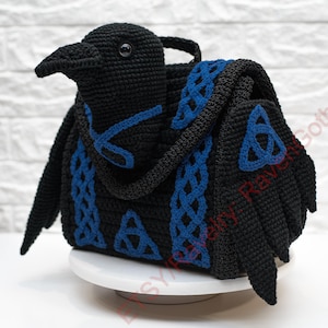 Op de afbeelding: Een zwarte gehaakte tas in de vorm van een raaf met blauwe Keltische knoopaccenten. De tas heeft een handvat en vleugels, en het ontwerp bevat blauwe randen en details. De kop van de raaf is gedetailleerd met een oog en een snavel.