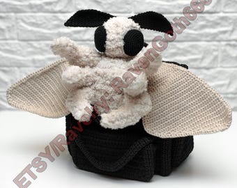 Könnte beinhalten: Handgefertigte Häkeltasche in Form einer Motte mit schwarzem Körper und Griff. Die Motte hat einen flauschigen cremefarbenen Körper, schwarze Augen und schwarze Fühler. Die Flügel sind hellbeige. Die Tasche steht auf einer weißen Oberfläche.