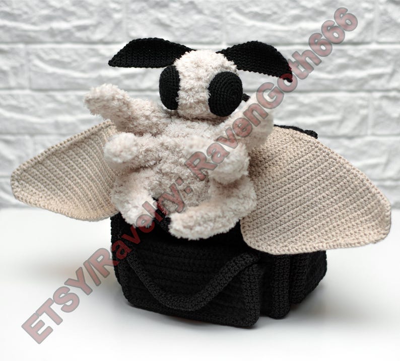 May include: A handmade crochet moth bag with a black body and handle. The moth has a fluffy cream-coloured body, black eyes, and black antennae. The wings are a light beige colour. The bag is sitting on a white surface.