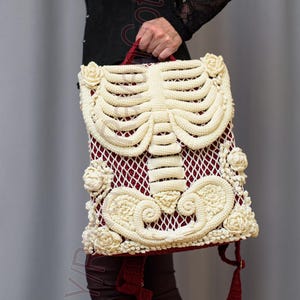 K&ouml;nnte beinhalten: Eine einzigartige Handtasche mit cremefarbenem H&auml;kelskelett-Design auf burgunderrotem Hintergrund. Die Tasche hat cremefarbene Rosen und einen roten Griff. Die Person tr&auml;gt schwarze Plateau-Stiefel.