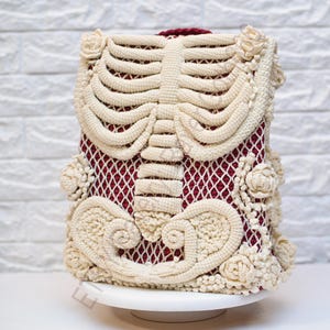 K&ouml;nnte beinhalten: Eine handgefertigte, mit H&auml;kelarbeit bedeckte Box mit einem cremefarbenen Skelettmuster auf einem bordeauxroten Netz-Hintergrund. Die H&auml;kelarbeit umfasst Brustkorb, Wirbels&auml;ule und florale Akzente. Die Box wird auf einem wei&szlig;en Kuchenst&auml;nder pr&auml;sentiert.