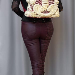 K&ouml;nnte beinhalten: Cremefarbener H&auml;kelrucksack mit Skelett-Rippen-Design und floralen Details, vor burgunderrotem Hintergrund. Die Person tr&auml;gt eine bordeauxfarbene Hose und schwarze Stiefel.