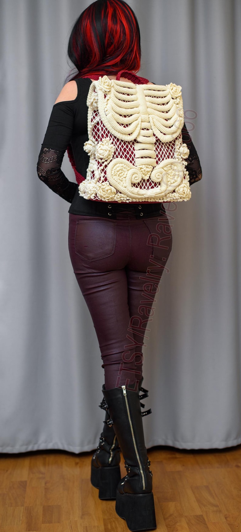 K&ouml;nnte beinhalten: Cremefarbener H&auml;kelrucksack mit Skelett-Rippen-Design und floralen Details, vor burgunderrotem Hintergrund. Die Person tr&auml;gt eine bordeauxfarbene Hose und schwarze Stiefel.