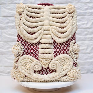 K&ouml;nnte beinhalten: Einzigartiger H&auml;kelrucksack mit cremefarbenem Brustkorb-Design auf bordeauxrotem Mesh-Hintergrund. Der Rucksack ist mit cremefarbenen floralen Akzenten und einem bordeauxroten Griff verziert. Die Tasche wird auf einer wei&szlig;en Tortenplatte pr&auml;sentiert.