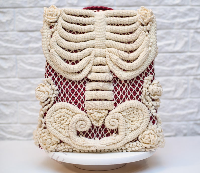 K&ouml;nnte beinhalten: Einzigartiger H&auml;kelrucksack mit cremefarbenem Brustkorb-Design auf bordeauxrotem Mesh-Hintergrund. Der Rucksack ist mit cremefarbenen floralen Akzenten und einem bordeauxroten Griff verziert. Die Tasche wird auf einer wei&szlig;en Tortenplatte pr&auml;sentiert.