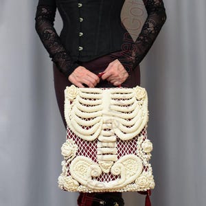 K&ouml;nnte beinhalten: Cremefarbene Makramee-Handtasche mit Skelett-Design und rotem Innenfutter. Die Tasche wird vor einer Person gehalten, die ein schwarzes Korsett, ein lang&auml;rmliges Oberteil und Plateaustiefel tr&auml;gt. Das Thema ist Gothic.