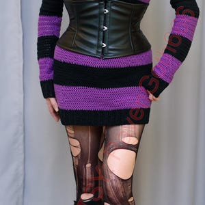 Pode incluir: Um vestido preto e roxo listrado com ombros de fora e um espartilho de couro preto. O traje inclui meias pretas rasgadas e botas de plataforma pretas. Um pingente de prata pende de um choker preto.