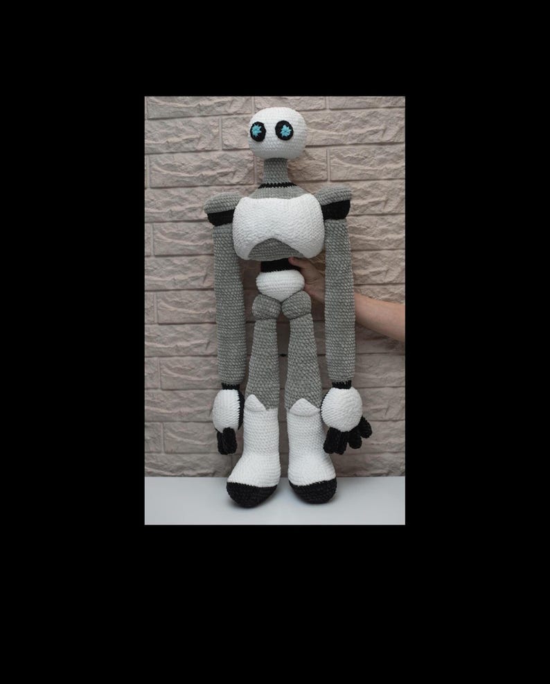 May include: A crocheted robot toy with gray and white yarn. The robot has a white head with black eyes and a gray body with white arms and legs. The robot is standing on a white surface.