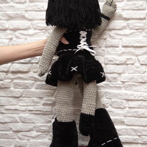 May include: A gray crocheted plush toy with black hair, a black skirt, a black corset with white lacing, and black boots. The toy is standing on a brown and black patterned surface.