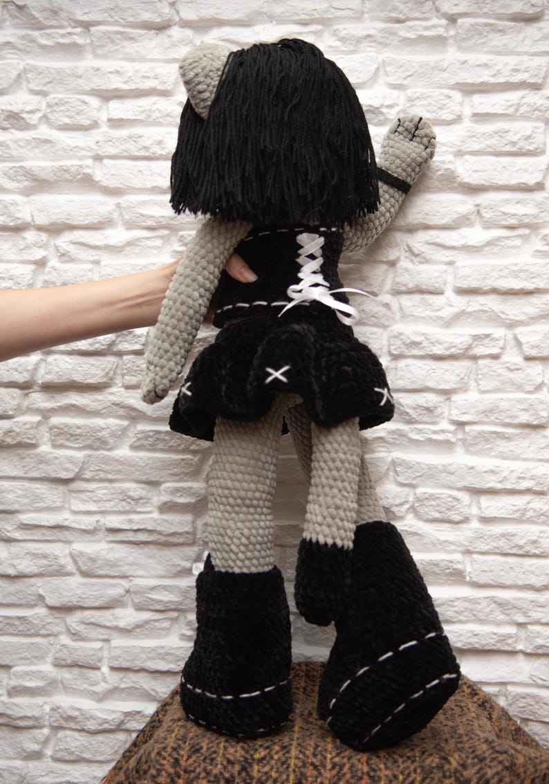 May include: A gray crocheted plush toy with black hair, a black skirt, a black corset with white lacing, and black boots. The toy is standing on a brown and black patterned surface.