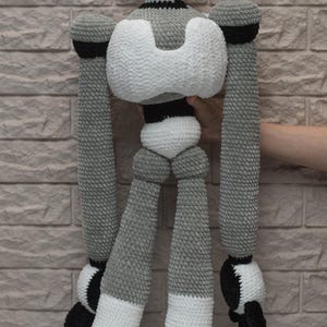 May include: A crocheted robot toy with a white head, gray body, and black accents. The robot has long arms and legs, and is standing on a white surface.