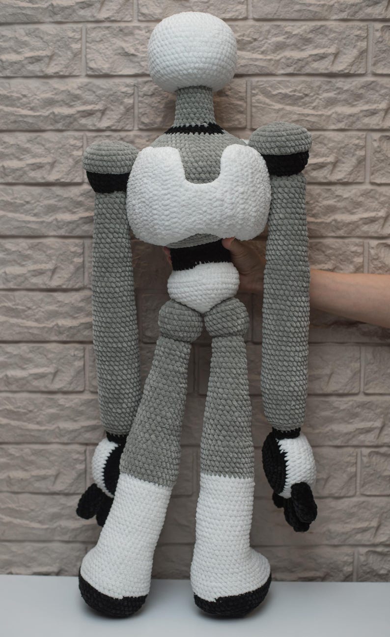 May include: A crocheted robot toy with a white head, gray body, and black accents. The robot has long arms and legs, and is standing on a white surface.