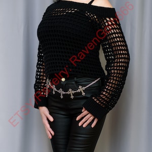 Op de afbeelding: Zwarte off-the-shoulder gehaakte trui met lange mouwen. Een zilveren kettingriem met kruisbedels wordt gedragen over een zwarte leren broek. Een zwarte choker-ketting met steraccenten maakt de look compleet. De outfit heeft een gotische esthetiek.