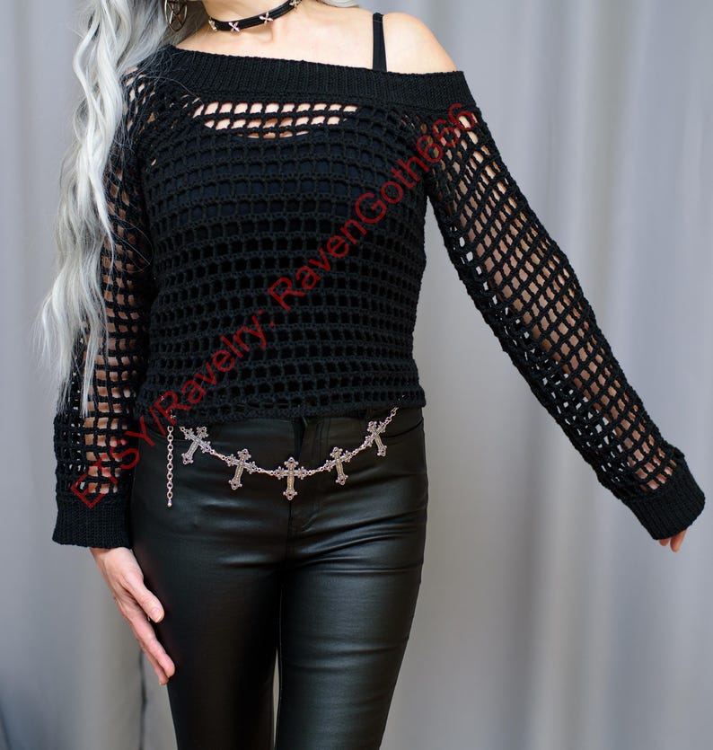 Op de afbeelding: Zwarte, off-the-shoulder, open geweven trui met lange mouwen. De trui heeft een uniek, open breisel ontwerp. Een zilveren kettingriem met kruisbedels wordt in de taille gedragen. De afbeelding is van een top in gotische stijl.