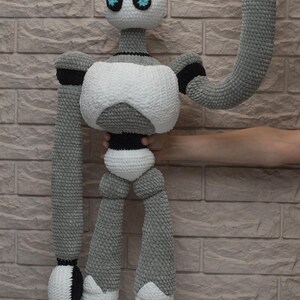 May include: A crocheted robot toy with a white body, gray limbs, and black accents. The robot has blue eyes and is waving with one arm.