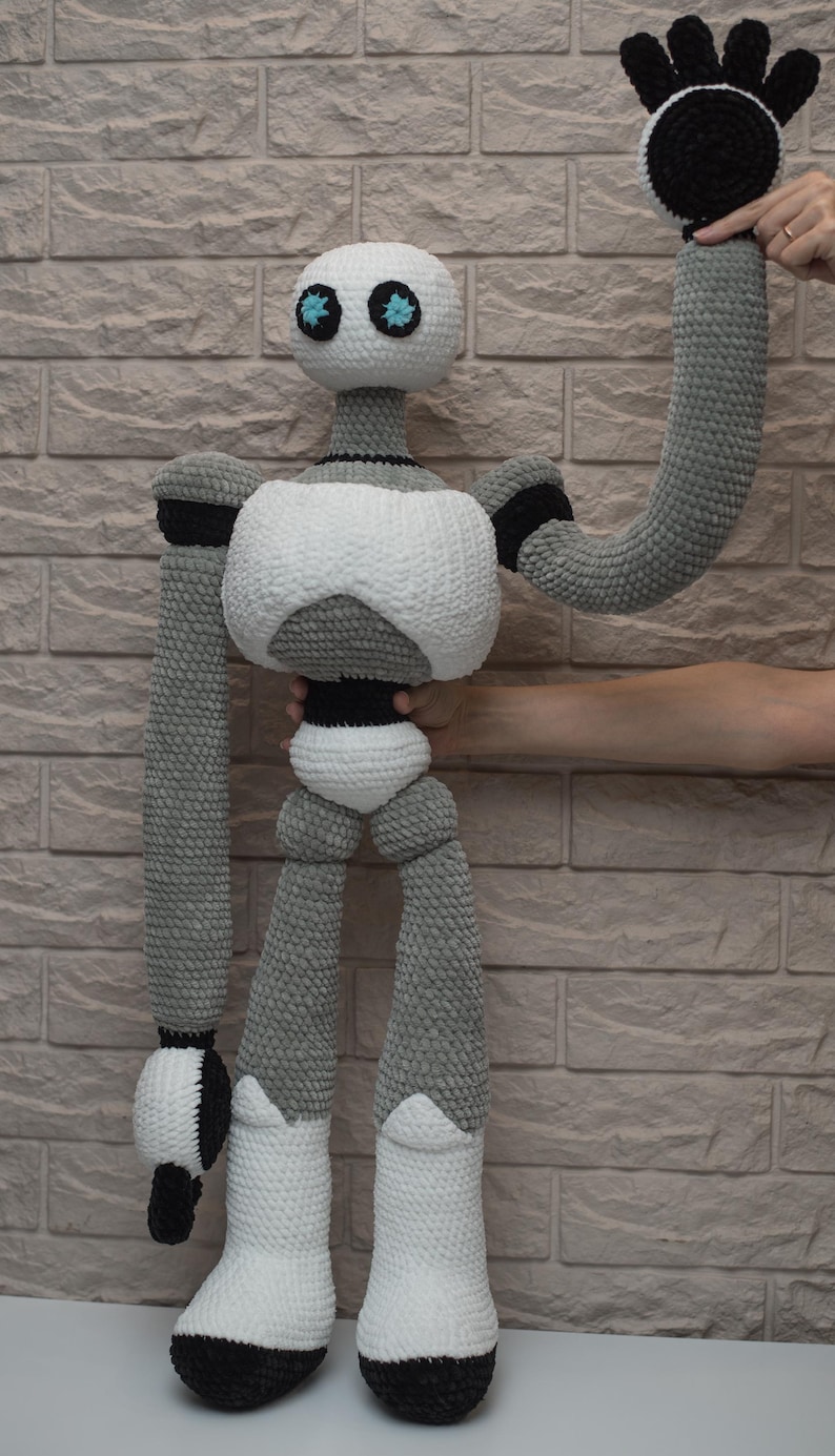 May include: A crocheted robot toy with a white body, gray limbs, and black accents. The robot has blue eyes and is waving with one arm.