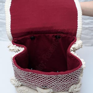 K&ouml;nnte beinhalten: Ge&ouml;ffnete Ansicht eines handgefertigten H&auml;kelrucksacks. Die Tasche hat eine cremefarbene, durchbrochene H&auml;kelau&szlig;enseite und ein weinrotes Innenfutter. Der Deckel wird mit einem silberfarbenen Verschluss gesichert. Das Design umfasst dekorative H&auml;kelelemente.