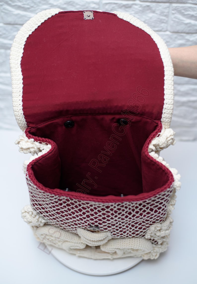 K&ouml;nnte beinhalten: Ge&ouml;ffnete Ansicht eines handgefertigten H&auml;kelrucksacks. Die Tasche hat eine cremefarbene, durchbrochene H&auml;kelau&szlig;enseite und ein weinrotes Innenfutter. Der Deckel wird mit einem silberfarbenen Verschluss gesichert. Das Design umfasst dekorative H&auml;kelelemente.