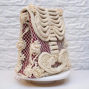 K&ouml;nnte beinhalten: Eine cremefarbene H&auml;keltasche mit Skelettmuster. Die Tasche hat ein weinrotes Innenfutter, das durch ein netzartiges Muster sichtbar ist. Cremefarbene H&auml;kelrosen verzieren die Seiten. Die Tasche steht auf einem wei&szlig;en Podest vor einer wei&szlig;en Backsteinmauer.