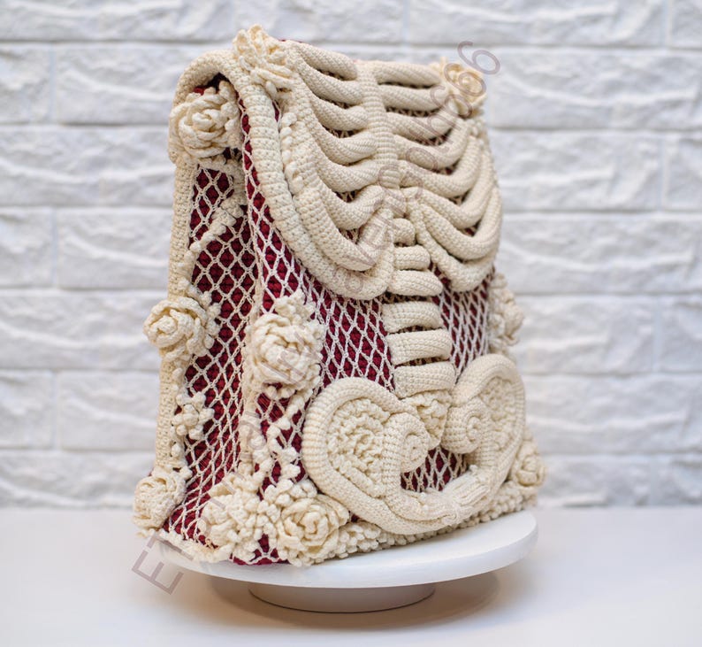 K&ouml;nnte beinhalten: Eine cremefarbene H&auml;keltasche mit Skelettmuster. Die Tasche hat ein weinrotes Innenfutter, das durch ein netzartiges Muster sichtbar ist. Cremefarbene H&auml;kelrosen verzieren die Seiten. Die Tasche steht auf einem wei&szlig;en Podest vor einer wei&szlig;en Backsteinmauer.