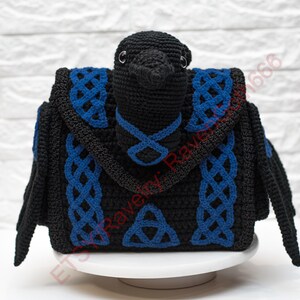 May include: A black crochet raven-shaped bag with blue Celtic knot accents. The bag features a bird-like head and wings, with a flap closure and a handle. The design includes a Celtic knot and a triangular symbol.