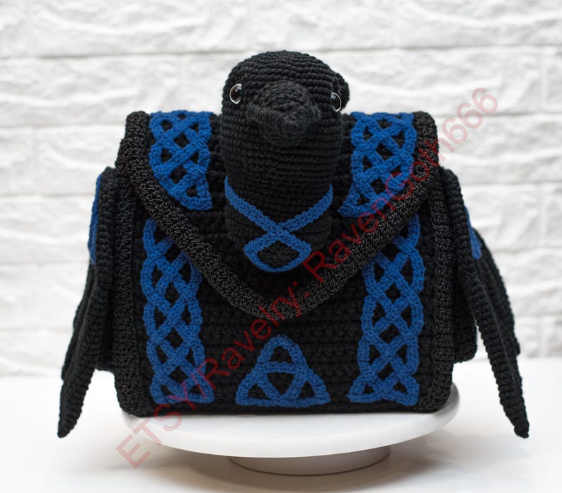 May include: A black crochet raven-shaped bag with blue Celtic knot accents. The bag features a bird-like head and wings, with a flap closure and a handle. The design includes a Celtic knot and a triangular symbol.