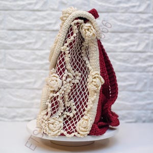 K&ouml;nnte beinhalten: Eine handgefertigte H&auml;keltasche in Creme, Burgunderrot und Wei&szlig;. Die Tasche hat ein cremefarbenes Blumendesign und Zierleisten mit einem burgunderroten Innenraum. Die Tasche wird auf einer wei&szlig;en Oberfl&auml;che vor einer wei&szlig;en Backsteinmauer pr&auml;sentiert.