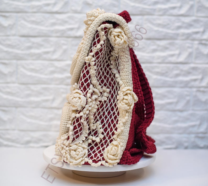 K&ouml;nnte beinhalten: Eine handgefertigte H&auml;keltasche in Creme, Burgunderrot und Wei&szlig;. Die Tasche hat ein cremefarbenes Blumendesign und Zierleisten mit einem burgunderroten Innenraum. Die Tasche wird auf einer wei&szlig;en Oberfl&auml;che vor einer wei&szlig;en Backsteinmauer pr&auml;sentiert.